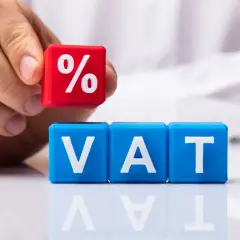 Ręka układa kostkę z symbolem procentu na kostkach z napisem VAT. Dowiedz się, jak zaksięgować czynsz za lokal z uwzględnieniem podatku.