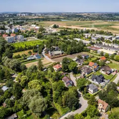 Osiedle domów jednorodzinnych na wzgórzach, otoczone zielenią i polami. W oddali widać zabudowania miejskie.