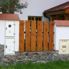 Zastanawiasz się, ile kosztuje ponowne podłączenie gazu do mieszkania w bloku? Na zdjęciu widzimy skrzynkę gazową z napisem "GAZ" przy kamiennym murku i drewnianym płocie.