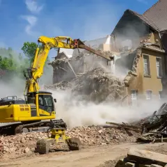 Żółta koparka burzy budynek, wzbijając tumany pyłu. Zastanawiasz się, ile kosztuje rozbiórka domu?