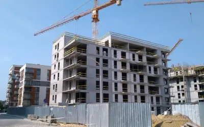 Budowa nowych mieszkań na sprzedaż w Bochni. Dwa bloki w budowie, jeden prawie gotowy, drugi w stanie surowym. Dźwigi budowlane na tle błękitnego nieba.