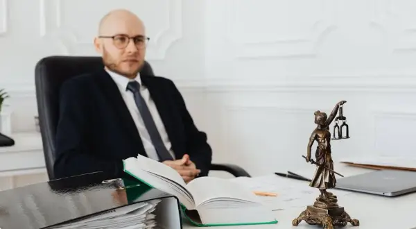 Prawnik w garniturze siedzi przy biurku, obok niego stos dokumentów i Statua Temidy. Zastanawiasz się, ile kosztuje notariusz przy zakupie mieszkania?