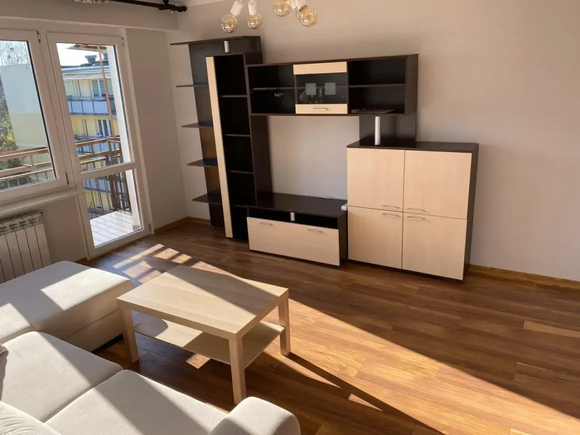 Jasne, nowoczesne mieszkania do wynajęcia Ostrołęka. Salon z meblościanką, stolikiem kawowym i kanapą. Duże okno z widokiem na balkon.