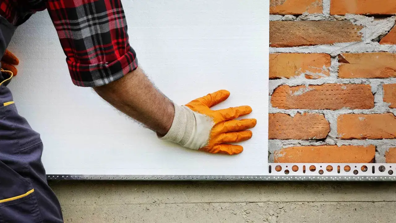 Ręka w pomarańczowej rękawicy przykłada biały styropian do ceglanej ściany. Dowiedz się, ile kosztuje termoizolacja budynku.