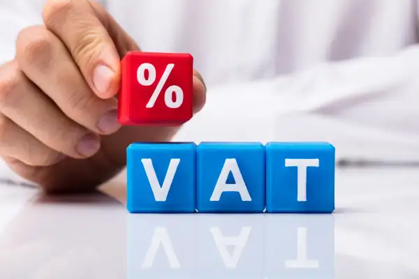 Ręka układa kostkę z symbolem procentu na kostkach z napisem VAT. Dowiedz się, jak zaksięgować czynsz za lokal z uwzględnieniem podatku.
